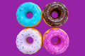 Four donuts arranged in a 2x2 grid on a white background, each with different frosting colors and sprinkles: blue, brown, light purple, and pink.